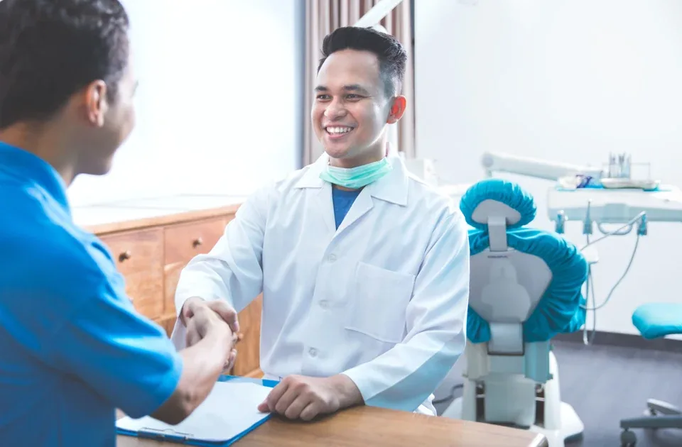 A dentist in a white coat shakes hands with a patient in a dental office.
