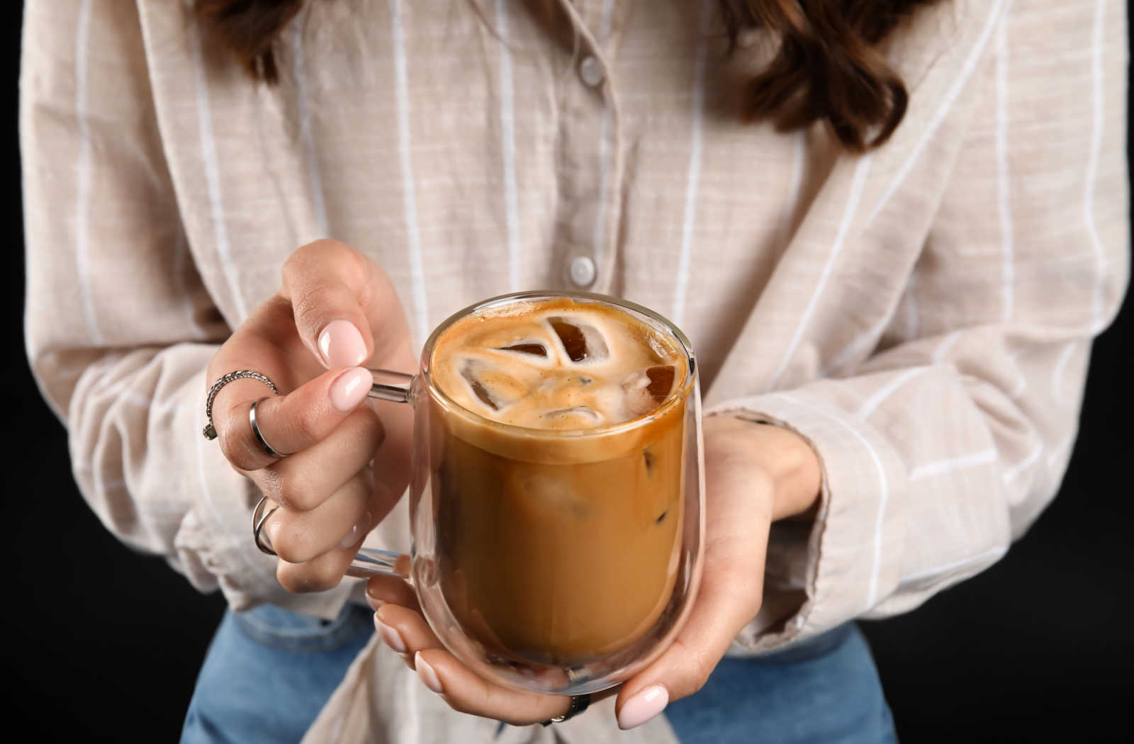 A close-up of a woman's hands holding a cup of iced coffee in a transparent glass cup.