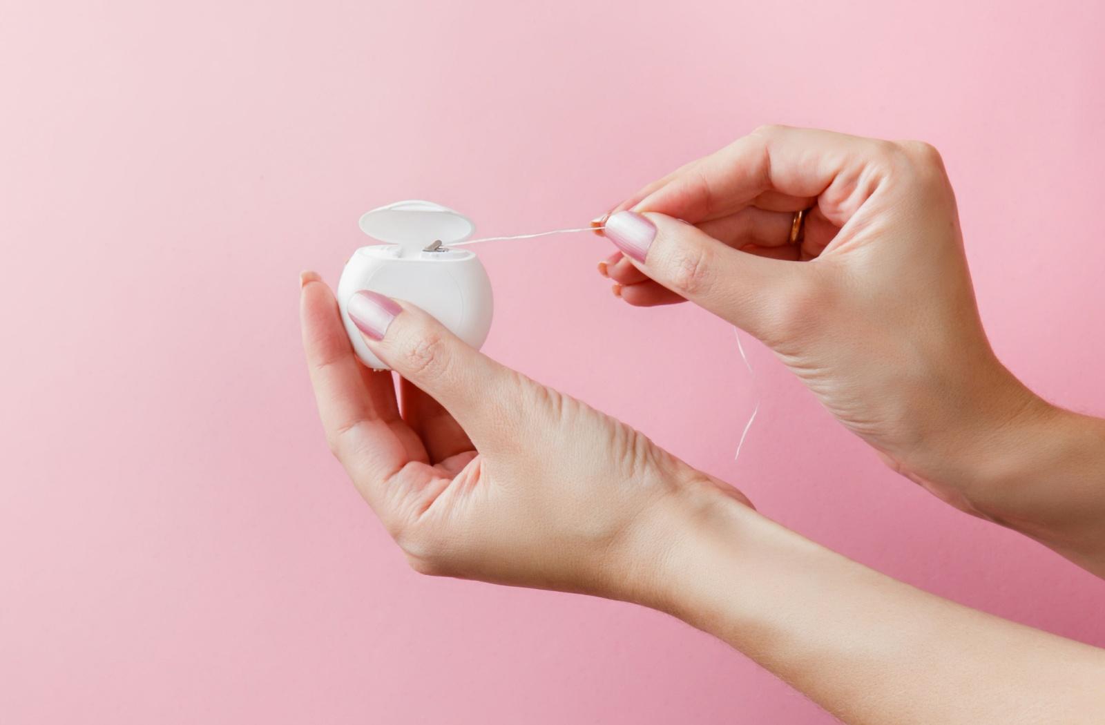 Close-up of hands pulling dental floss from a container, representing traditional flossing for plaque removal and gum health.