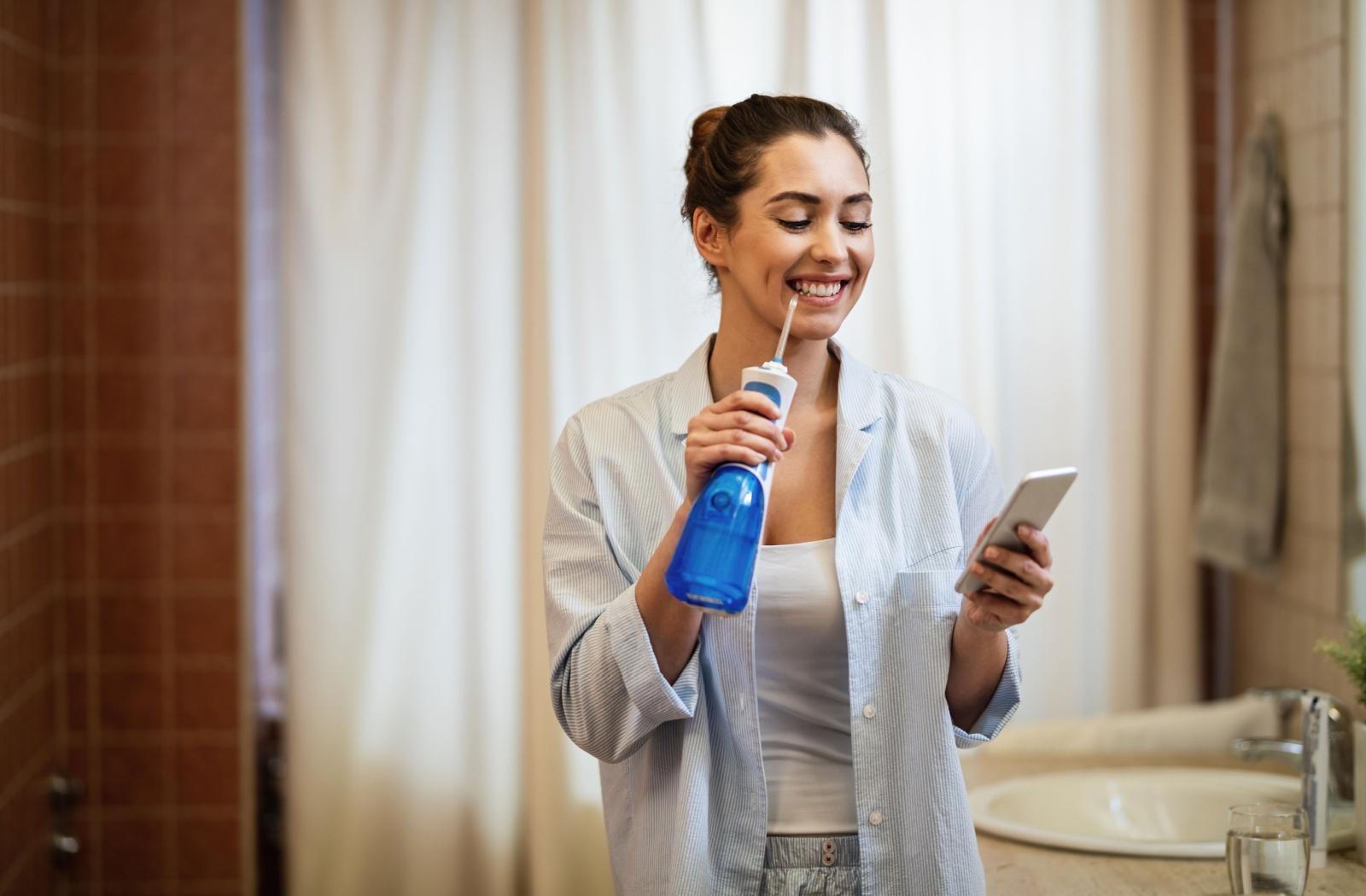 Woman using a water flosser in the bathroom while looking at her phone, highlighting Waterpik use for daily oral hygiene.