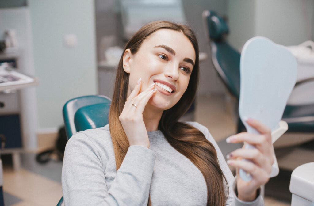 A woman sitting in a dentist’s chair and smiling into a handheld mirror