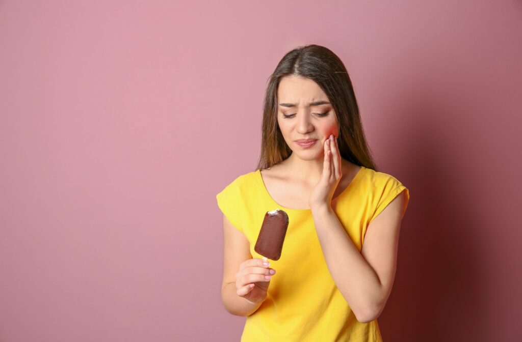 A woman in a yellow shirt standing against a pink background and holding an ice cream bar in one hand as she holds her cheek in pain with her other hand