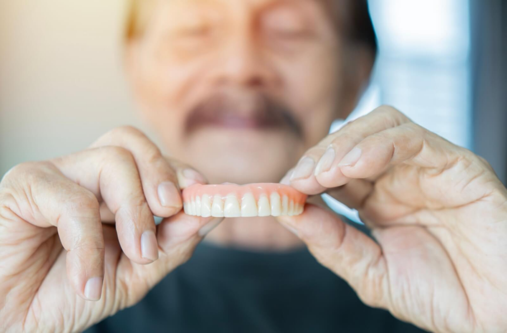 An older adult holding up upper dentures. Face is blurry but hands and denture are in focus.