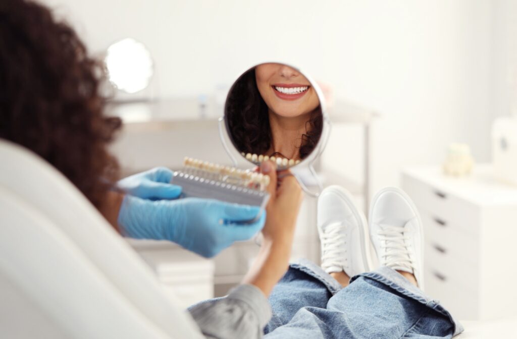 Close up photo of a dental patient smiling in hand-held mirror while a dentist selects veneer shades for their teeth